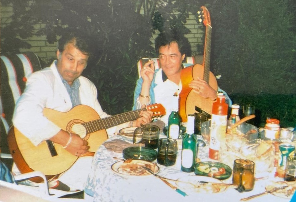 Johan Heeren speelt gitaar en rechts van hem zit Jack Jersey met sigaret en gitaar naar hem te kijken. Ze zitten op tuinstoelen aan een tafel vol hapjes en drankjes