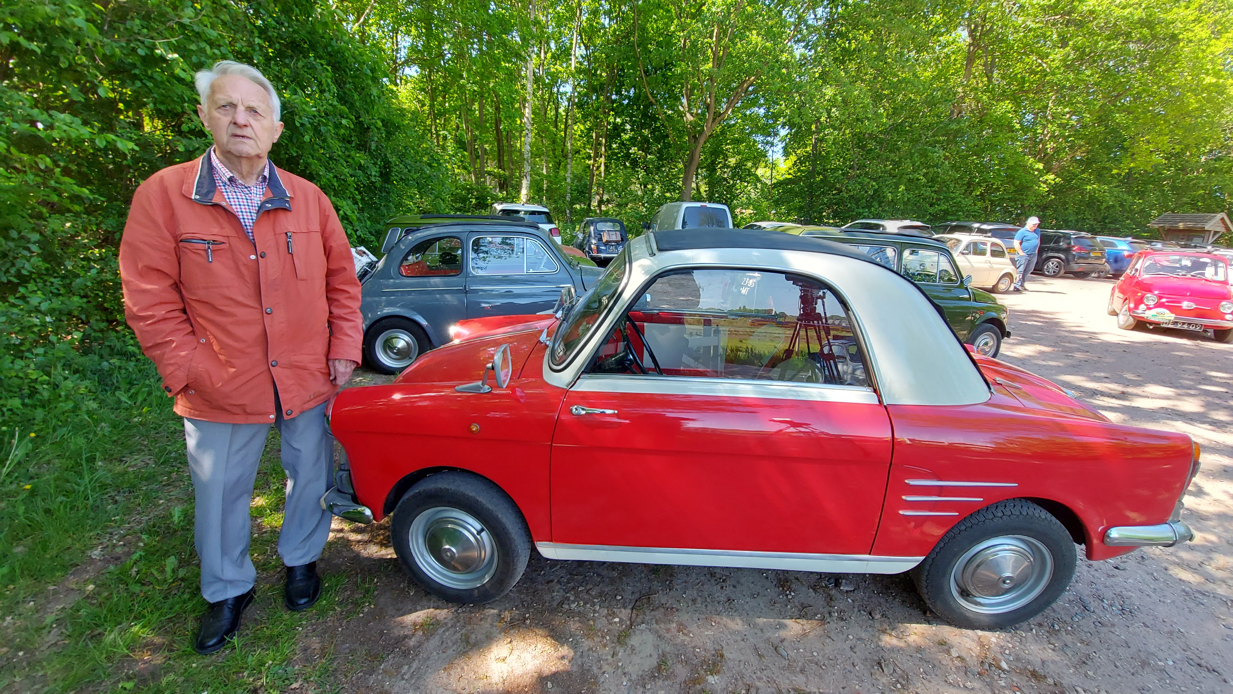 Ad Kostermans in grijze broek en rode jas poseert bij zijn Autobianchi S, een vuurrode auto met witte kap